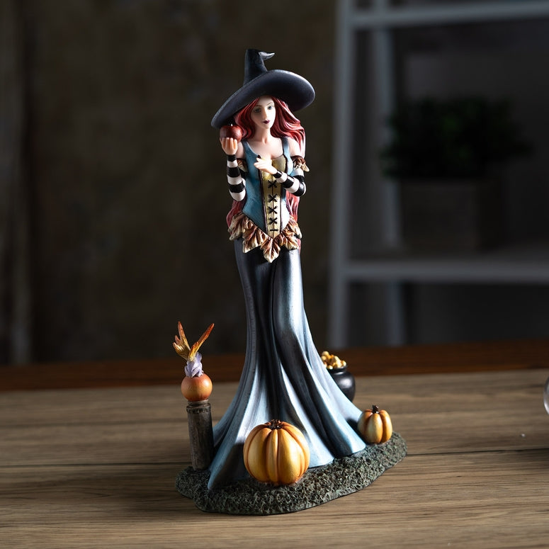 A Halloween witch figurine standing on a textured base with small pumpkins, set on a wooden tabletop with a blurred indoor background.