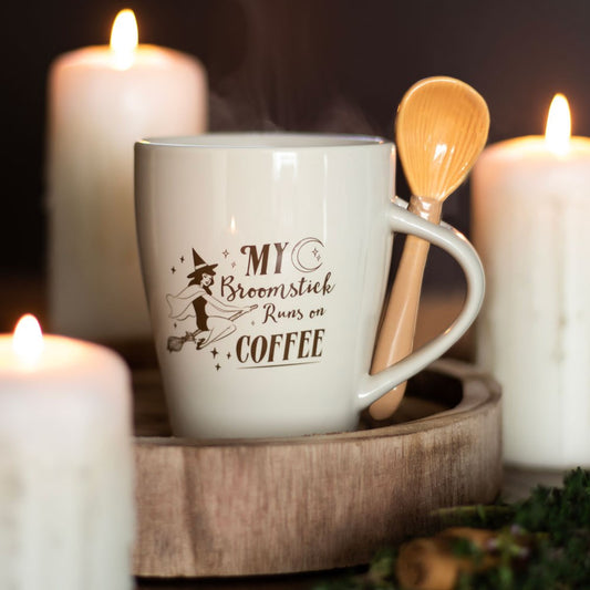 White mug with 'My Broomstick Runs on Coffee' text, wooden spoon, and candles on a dark background