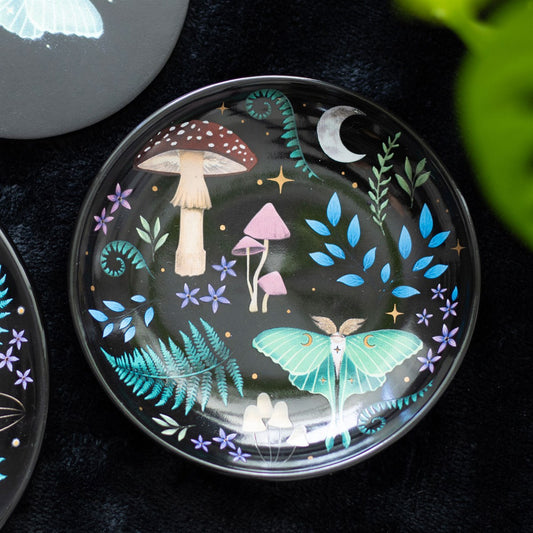 A round black trinket dish with a mystical forest design, featuring colorful plants, mushrooms, and an insect.