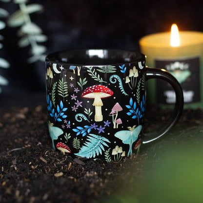 A black ceramic mug with an all-over mystical forest print, featuring mushrooms, leaves, and whimsical characters, placed on a bed of soil with a candle lit in the background.