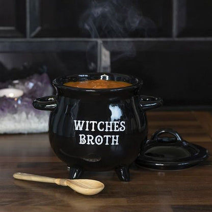 A black ceramic soup bowl designed to resemble a witch's cauldron with 'WITCHES BROTH' text on it, accompanied by a wooden spoon, on a kitchen counter.