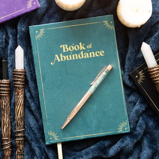 Green book titled 'Book of Abundance' with a pen on a dark fabric background