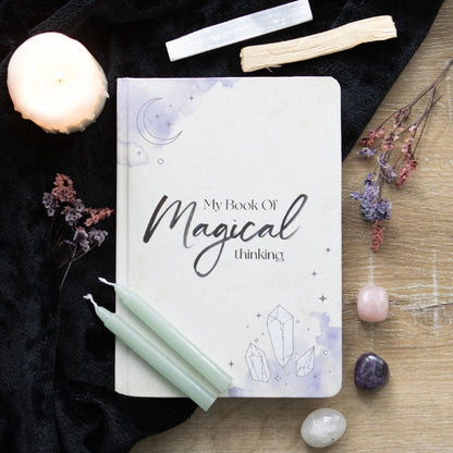 Notebook titled 'My Book Of Magical Thinking' surrounded by candles, crystals, and flowers on a wooden surface.