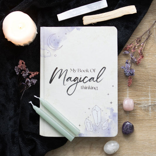 Notebook titled 'My Book Of Magical Thinking' surrounded by candles, crystals, and flowers on a wooden surface.